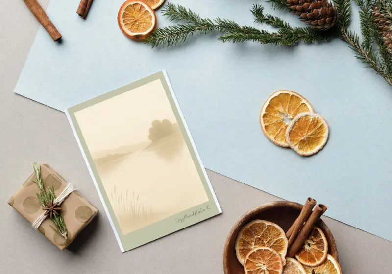 Notebook with a pen on a table with decorative items like dried oranges and cinnamon sticks. Still Water Printable Greeting Card.